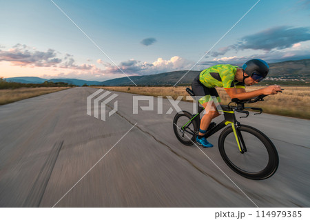 Triathlete riding his bicycle during sunset, preparing for a marathon. The warm colors of the sky provide a beautiful backdrop for his determined and focused effort. 114979385