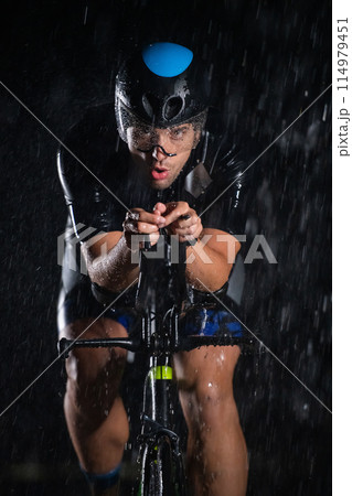 A triathlete braving the rain as he cycles through the night, preparing himself for the upcoming marathon. The blurred raindrops in the foreground and the dark, moody atmosphere in the background add A triathlete braving the rain as he cycles through the night, preparing himself for the upcoming marathon. The blurred raindrops in the foreground and the dark, moody atmosphere in the background add 114979451