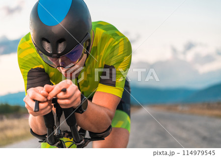 Close up photo of triathlete riding his bicycle during sunset, preparing for a marathon. The warm colors of the sky provide a beautiful backdrop for his determined and focused effort. Close up photo of triathlete riding his bicycle during sunset, preparing for a marathon. The warm colors of the sky provide a beautiful backdrop for his determined and focused effort. 114979545