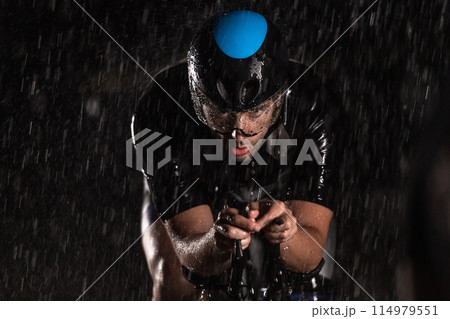 A triathlete braving the rain as he cycles through the night, preparing himself for the upcoming marathon. The blurred raindrops in the foreground and the dark, moody atmosphere in the background add A triathlete braving the rain as he cycles through the night, preparing himself for the upcoming marathon. The blurred raindrops in the foreground and the dark, moody atmosphere in the background add 114979551
