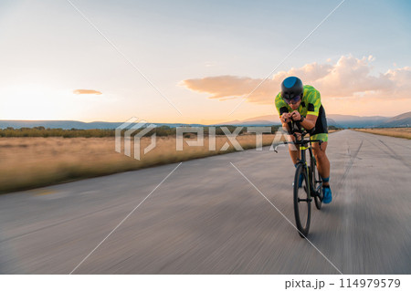 Triathlete riding his bicycle during sunset, preparing for a marathon. The warm colors of the sky provide a beautiful backdrop for his determined and focused effort. Triathlete riding his bicycle during sunset, preparing for a marathon. The warm colors of the sky provide a beautiful backdrop for his determined and focused effort. 114979579