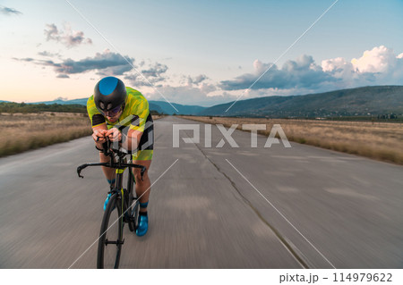Triathlete riding his bicycle during sunset, preparing for a marathon. The warm colors of the sky provide a beautiful backdrop for his determined and focused effort. 114979622
