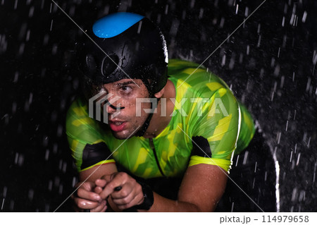A triathlete braving the rain as he cycles through the night, preparing himself for the upcoming marathon. The blurred raindrops in the foreground and the dark, moody atmosphere in the background add A triathlete braving the rain as he cycles through the night, preparing himself for the upcoming marathon. The blurred raindrops in the foreground and the dark, moody atmosphere in the background add 114979658