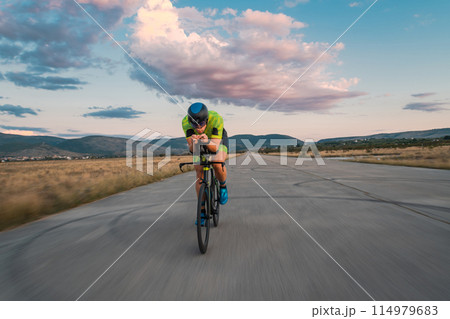 Triathlete riding his bicycle during sunset, preparing for a marathon. The warm colors of the sky provide a beautiful backdrop for his determined and focused effort. 114979683
