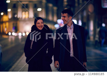 Happy multicultural business couple walking together outdoors in an urban city street at night near a jewelry shopping store window. 114980185
