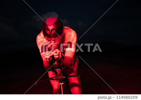 A triathlete rides his bike in the darkness of night, pushing himself to prepare for a marathon. The contrast between the darkness and the light of his bike creates a sense of drama and highlights the A triathlete rides his bike in the darkness of night, pushing himself to prepare for a marathon. The contrast between the darkness and the light of his bike creates a sense of drama and highlights the 114980209