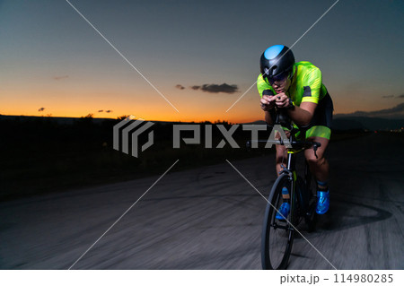 A triathlete rides his bike in the darkness of night, pushing himself to prepare for a marathon. The contrast between the darkness and the light of his bike creates a sense of drama and highlights the 114980285