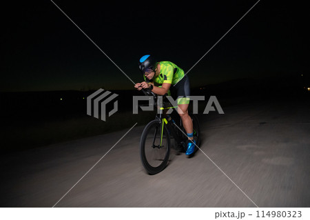 A triathlete rides his bike in the darkness of night, pushing himself to prepare for a marathon. The contrast between the darkness and the light of his bike creates a sense of drama and highlights the A triathlete rides his bike in the darkness of night, pushing himself to prepare for a marathon. The contrast between the darkness and the light of his bike creates a sense of drama and highlights the 114980323