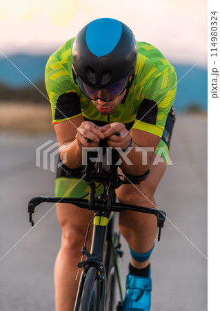 Close up photo of triathlete riding his bicycle during sunset, preparing for a marathon. The warm colors of the sky provide a beautiful backdrop for his determined and focused effort. 114980324