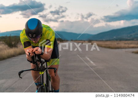 Triathlete riding his bicycle during sunset, preparing for a marathon. The warm colors of the sky provide a beautiful backdrop for his determined and focused effort. 114980474