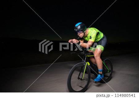 A triathlete rides his bike in the darkness of night, pushing himself to prepare for a marathon. The contrast between the darkness and the light of his bike creates a sense of drama and highlights the 114980541