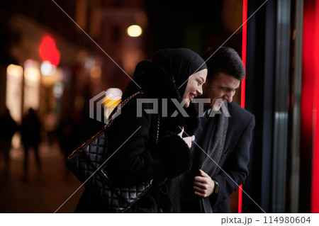 Happy multicultural business couple walking together outdoors in an urban city street at night near a jewelry shopping store window. Happy multicultural business couple walking together outdoors in an urban city street at night near a jewelry shopping store window. 114980604