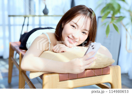 部屋の中で椅子に寝そべってスマホを見る女性 114980653