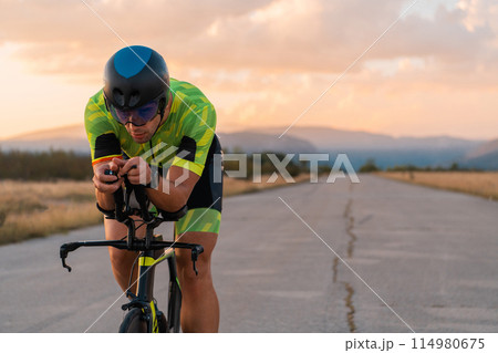 Triathlete riding his bicycle during sunset, preparing for a marathon. The warm colors of the sky provide a beautiful backdrop for his determined and focused effort. Triathlete riding his bicycle during sunset, preparing for a marathon. The warm colors of the sky provide a beautiful backdrop for his determined and focused effort. 114980675