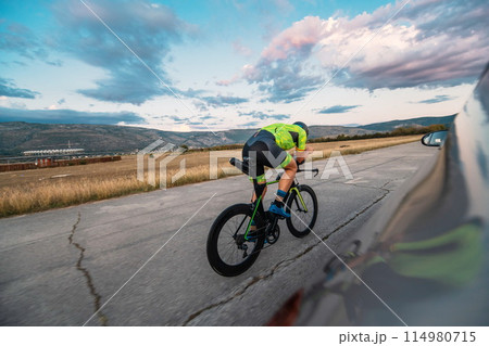 Triathlete riding his bicycle during sunset, preparing for a marathon. The warm colors of the sky provide a beautiful backdrop for his determined and focused effort. 114980715