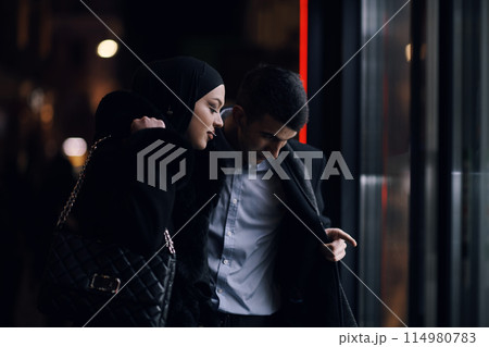 Happy multicultural business couple walking together outdoors in an urban city street at night near a jewelry shopping store window. 114980783