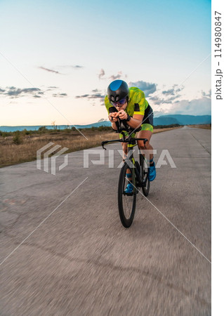 Triathlete riding his bicycle during sunset, preparing for a marathon. The warm colors of the sky provide a beautiful backdrop for his determined and focused effort. Triathlete riding his bicycle during sunset, preparing for a marathon. The warm colors of the sky provide a beautiful backdrop for his determined and focused effort. 114980847