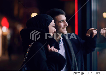 Happy multicultural business couple walking together outdoors in an urban city street at night near a jewelry shopping store window. Happy multicultural business couple walking together outdoors in an urban city street at night near a jewelry shopping store window. 114980930