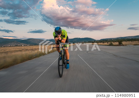 Triathlete riding his bicycle during sunset, preparing for a marathon. The warm colors of the sky provide a beautiful backdrop for his determined and focused effort. 114980951
