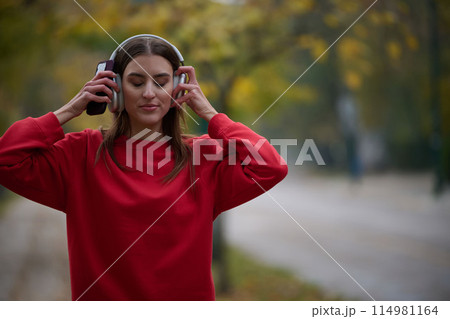 Portrait of running woman after jogging in the park on autumn seasson. Female fitness model training outside on a cozy fall day and listening to music over smartphone. 114981164