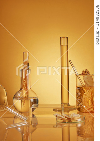 Yellow background photo with frontal shot has ginseng composition and lab instruments including measuring cylinder, boiling flask, beaker, petri dish and a blank glass showcase podium in center Yellow background photo with frontal shot has ginseng composition and lab instruments including measuring cylinder, boiling flask, beaker, petri dish and a blank glass showcase podium in center 114982536