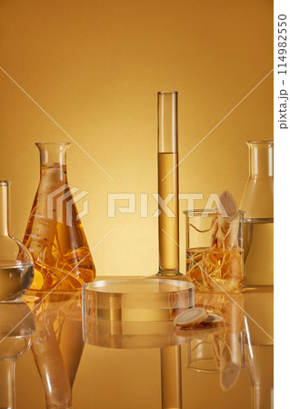 Yellow color tone photo with ginseng experiment theme, glassware in lab containing yellow liquid and ginseng root decorated behind a round glass pedestal. Frontal shot photo with space for text Yellow color tone photo with ginseng experiment theme, glassware in lab containing yellow liquid and ginseng root decorated behind a round glass pedestal. Frontal shot photo with space for text 114982550