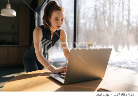 Focused sporty young woman in outfit using laptop standing at desk by window on sunny day, thinking of online creative task, browsing internet, search information, working on computer typing email 114988418