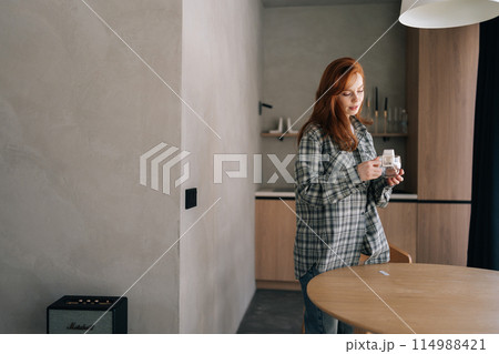 Attractive young woman putting bag of drip coffee in glass cup at table in kitchen room. Pretty red-haired female opening coffee drip bag at home standing by table in daytime. 114988421