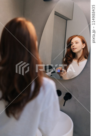 Vertical shot of young woman playing with long red hair looking in mirror standing in bathroom in morning. Reflection on mirror of pretty redhead female preens, admires, straightening hairstyle 114988435