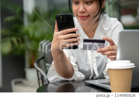 A young Asian woman holding a credit card and her smartphone while sitting at a table outdoors. A young Asian woman holding a credit card and her smartphone while sitting at a table outdoors. 114990826