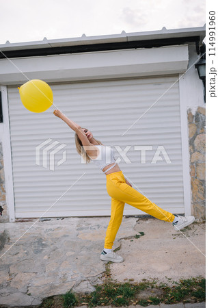 Cheerful young woman wearing yellow pants walking with yellow balloon on the street Cheerful young woman wearing yellow pants walking with yellow balloon on the street 114991960
