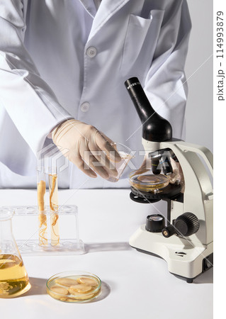 Frontal shot of a scientist holding a glass beaker containing yellow liquid, preparing to pour it into a petri dish on a microscope, which placed on white countertop next to ginseng and lab tools Frontal shot of a scientist holding a glass beaker containing yellow liquid, preparing to pour it into a petri dish on a microscope, which placed on white countertop next to ginseng and lab tools 114993879