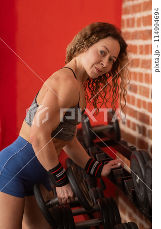 Middle aged woman takes dumbbell from rack in gym. Vertical photo.  114994694