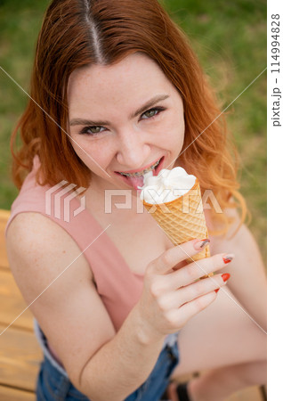 Young beautiful red-haired woman smiling with braces and eating an ice cream cone outdoors. Vertical photo. Young beautiful red-haired woman smiling with braces and eating an ice cream cone outdoors. Vertical photo. 114994828