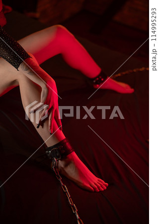 A faceless woman sits on a bed with her ankles handcuffed. BDSM concept. Vertical photo.  114995293