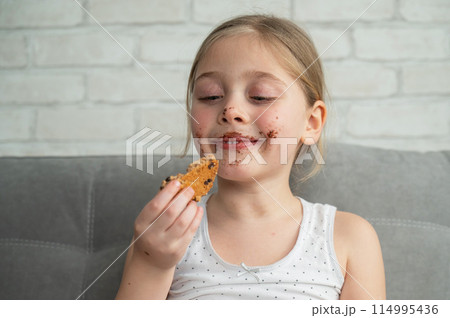 A cute little girl covered in chocolate eats cookies while sitting on the sofa. 114995436