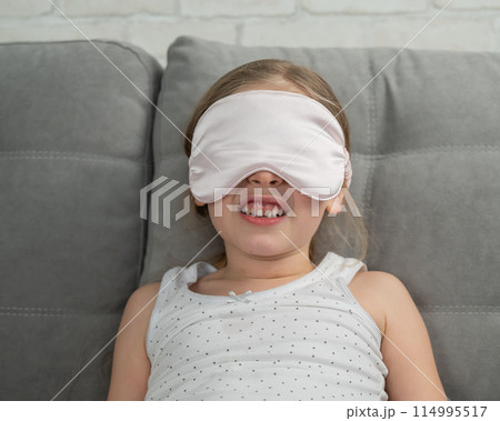 Little Caucasian girl puts on a sleep mask while sitting on the sofa. Doesn't want to sleep.  114995517