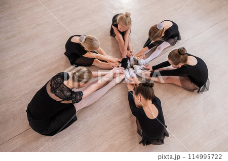Caucasian woman and five little girls sit in a circle and do stretching at a ballet school.  114995722