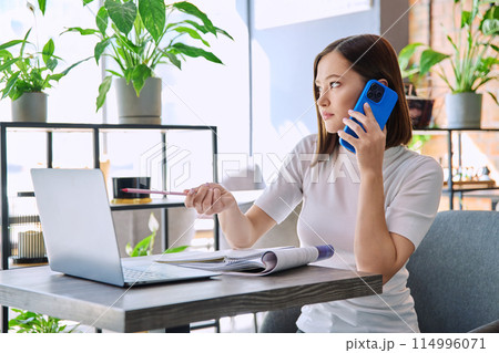 Young woman sitting at table with laptop talking on mobile phone in coworking 114996071