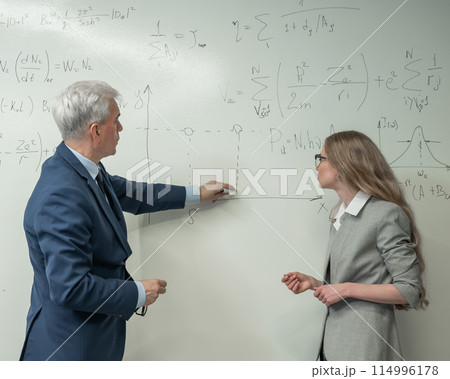 An elderly professor explains a subject to a student at a white board. An elderly professor explains a subject to a student at a white board. 114996178