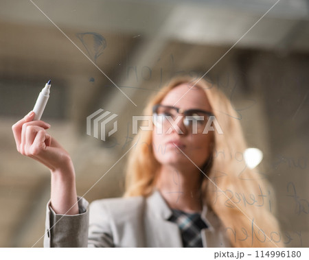 Caucasian woman with glasses writes text in English on a glass wall.  114996180
