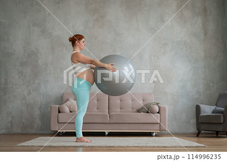 Pregnant red-haired woman doing arm exercises with fitball at home. 114996235