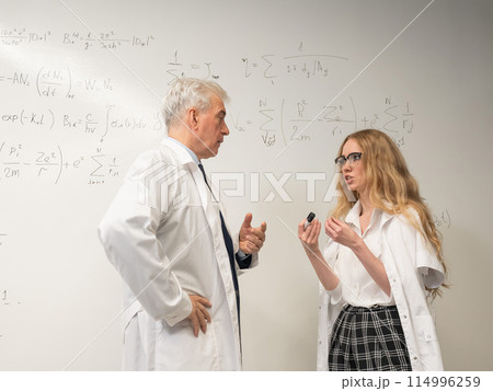An elderly man and a young woman at a white board. Colleagues scientists discuss work issues.  114996259