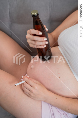 Faceless pregnant woman drinks beer while sitting on the sofa.  114996595