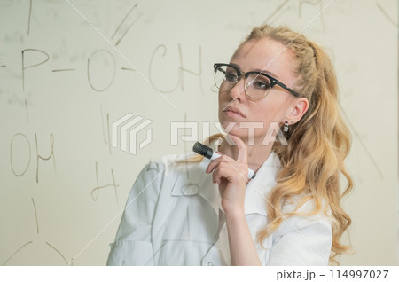 A Caucasian woman in a medical gown thinks and finalizes formulas on a transparent wall. A Caucasian woman in a medical gown thinks and finalizes formulas on a transparent wall. 114997027