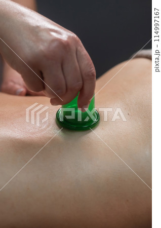 A woman undergoes an anti-cellulite massage procedure using a vacuum jar. Close-up of the lower back. Vertical photo.  114997167