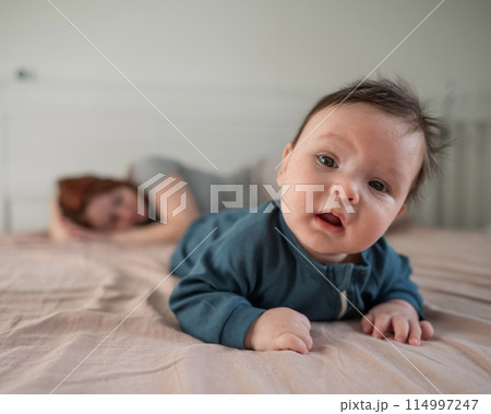 A three-month-old boy lies on his stomach on the bed and his mother sleeps behind him. Postpartum depression. A three-month-old boy lies on his stomach on the bed and his mother sleeps behind him. Postpartum depression. 114997247