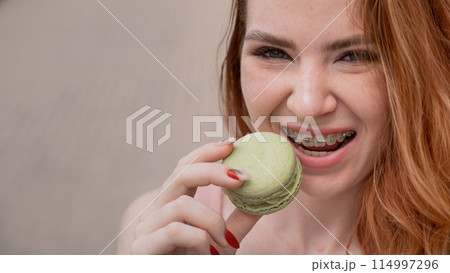 Young red-haired woman with braces eating macaron cake.  114997296