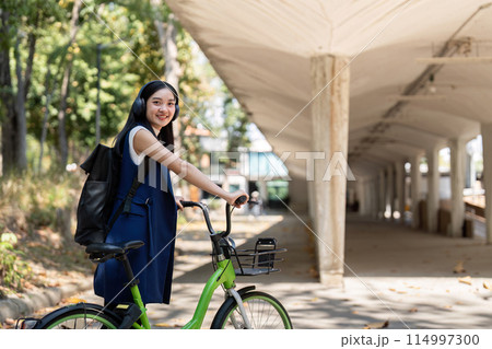 Businesswoman commuting by bicycle with headphones in urban park. Concept of eco friendly transport, active lifestyle, and outdoor fitness Businesswoman commuting by bicycle with headphones in urban park. Concept of eco friendly transport, active lifestyle, and outdoor fitness 114997300