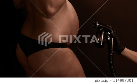 Close-up of the belly of a pregnant woman undergoing an instant tanning procedure. The master applies bronzer with a spray. 114997301